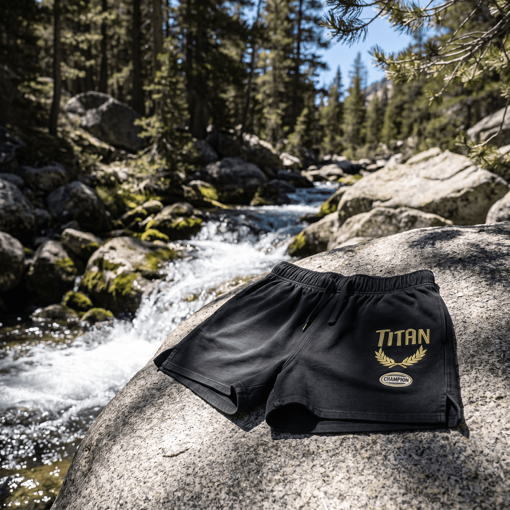 Black athletic shorts with "TITAN" and a gold laurel graphic, featuring a drawstring waist on a rocky streamside.