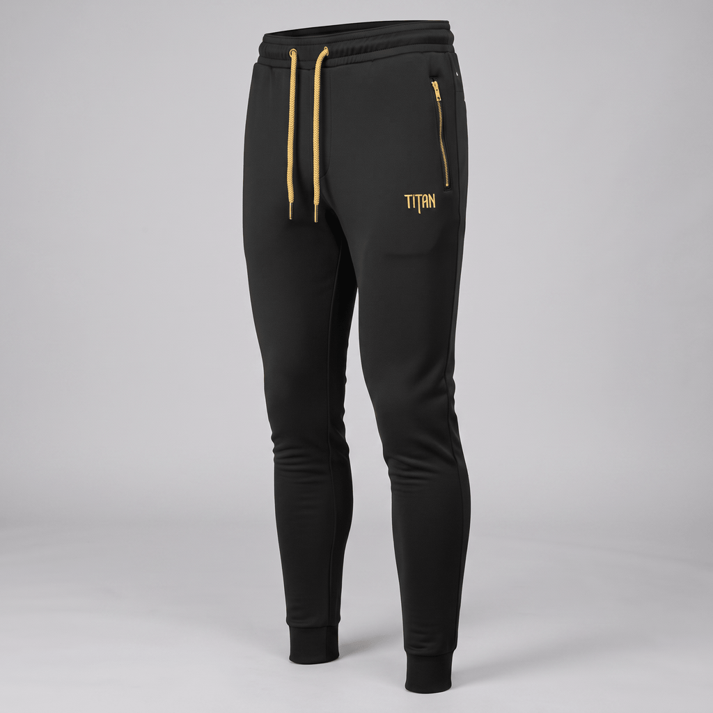Black athletic joggers with gold drawstring, zipper pocket, and Titan logo against a grey studio backdrop.