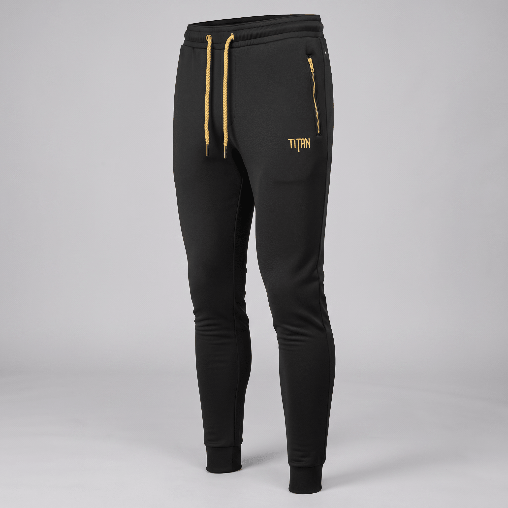 Black athletic joggers with gold drawstring, zipper pocket, and Titan logo against a grey studio backdrop.