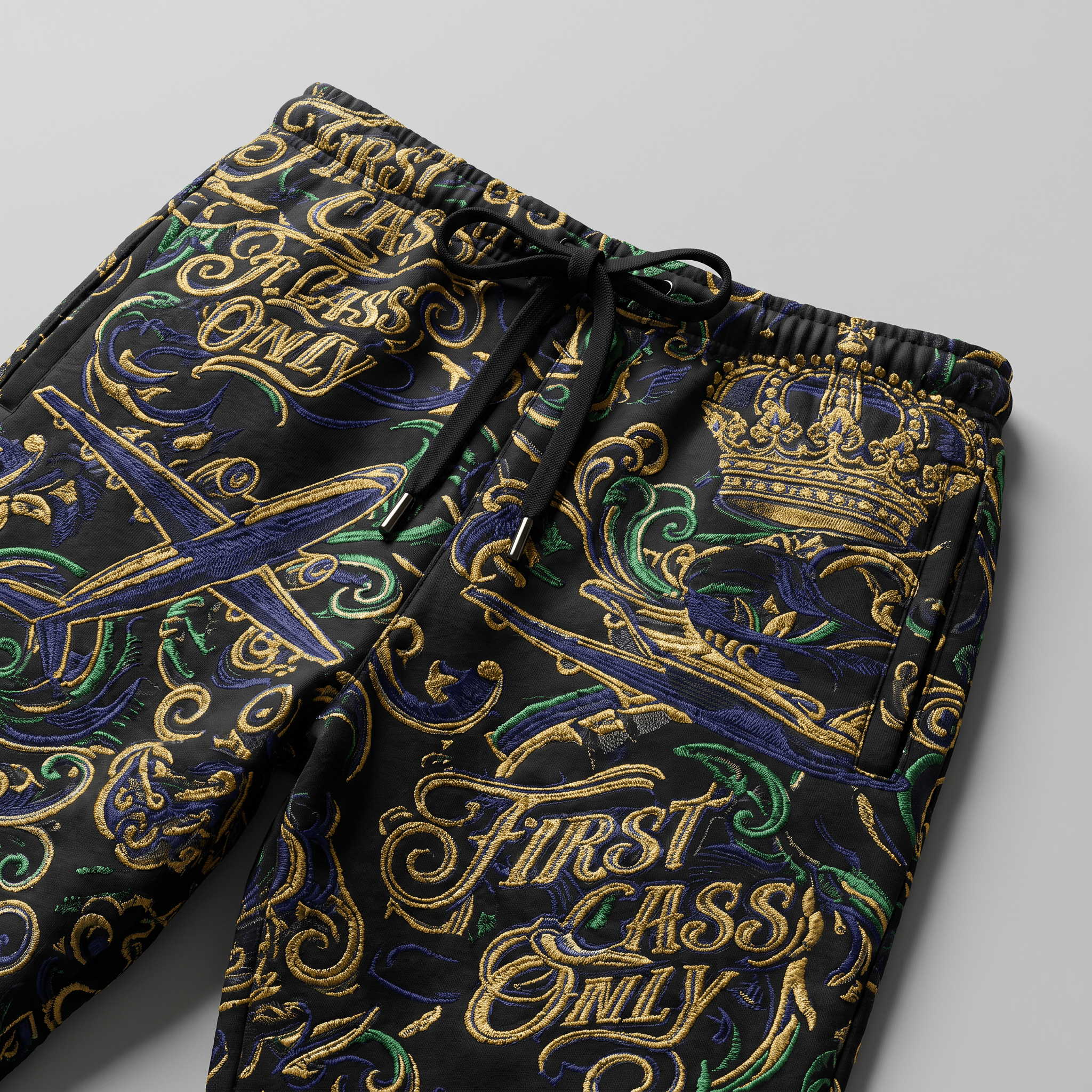 Black sweatpants with ornate gold, blue, and green embroidery featuring crowns, airplanes, and First Class Only text.
