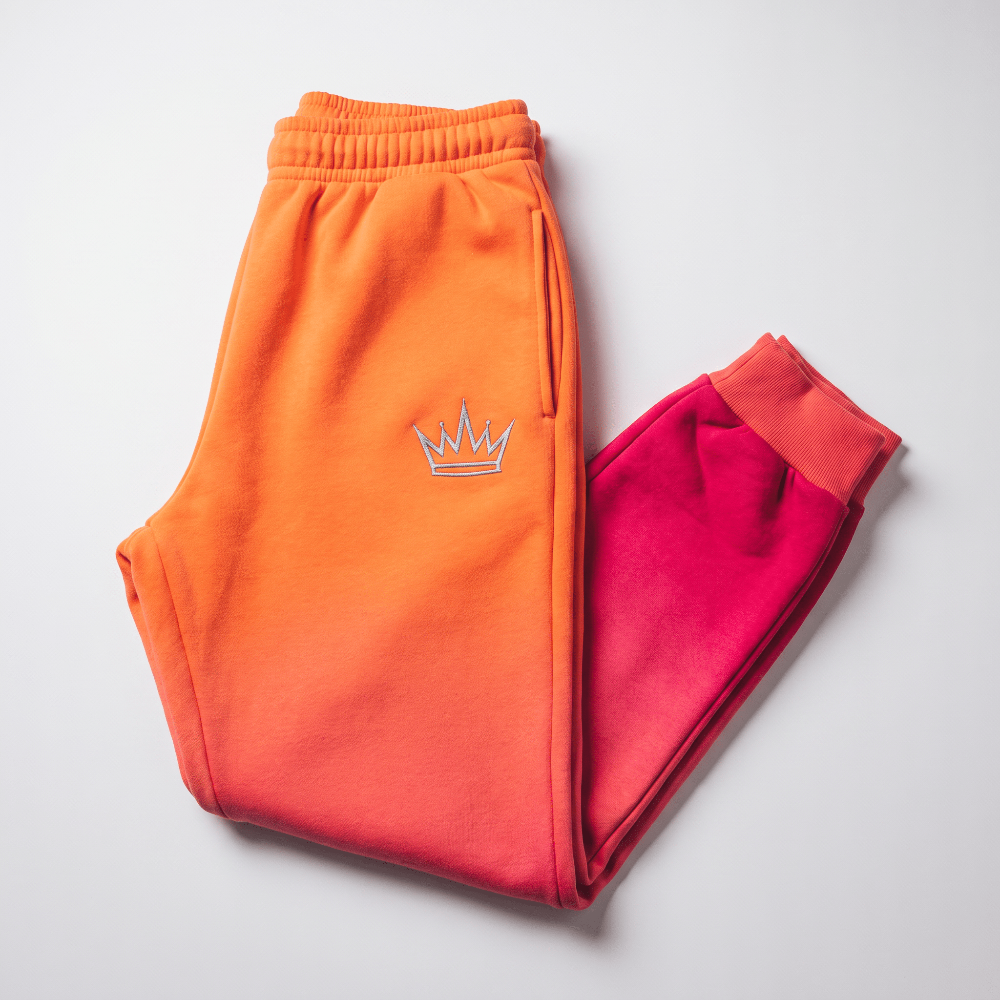 Orange and pink ombre sweatpants with embroidered silver crown logo on light background.