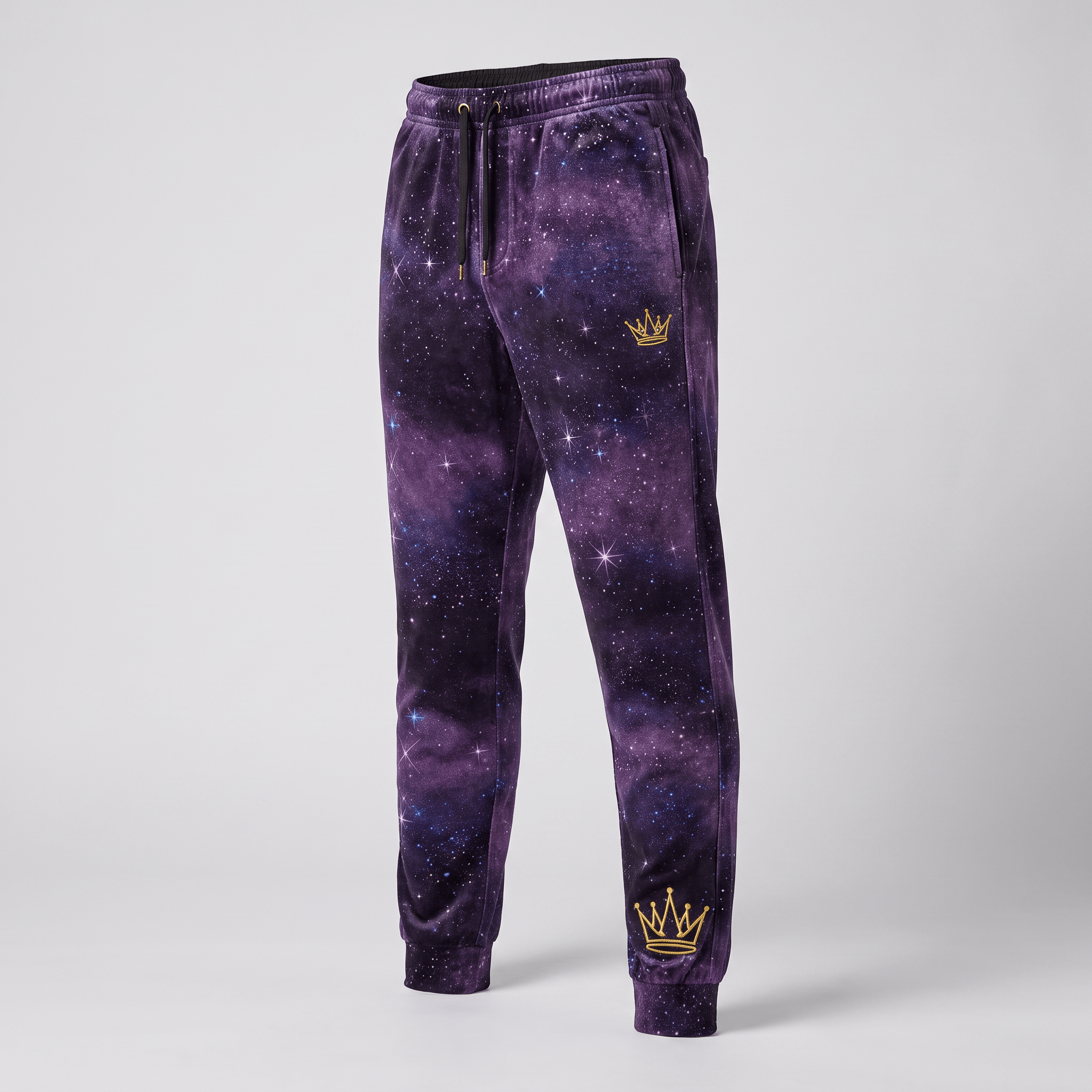Purple galaxy print joggers with gold crown embroidery, drawstring waist, and cuffed ankles on a neutral background.