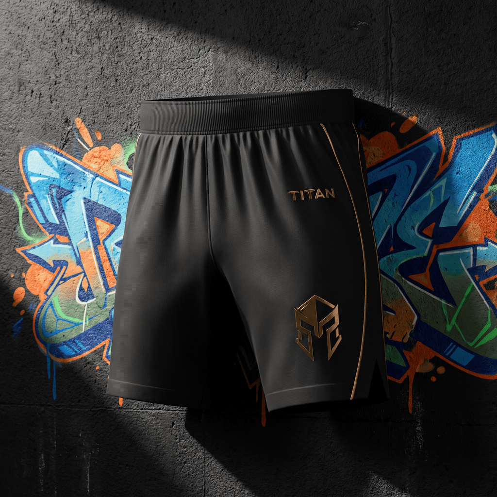 Black athletic shorts featuring gold "TITAN" text, a gold geometric logo, and gold piping against a graffiti wall.