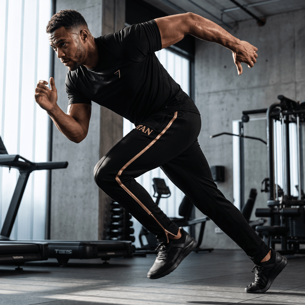 Male athlete sprinting in a gym wearing black TITAN track pants with gold stripes and matching black t-shirt.