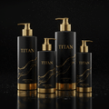 Four matte black TITAN shower gel bottles with gold pumps, bases, and gold swirl accents on a wet reflective surface.