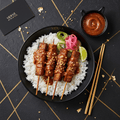 Tempeh satay skewers with peanut sauce and white rice in a black bowl, with pickled onions, lime, and gold chopsticks.