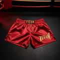Red satin boxing shorts with gold TITAN embroidery, elastic waistband, and drawstring on a black floor.