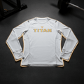 White long-sleeve compression shirt with gold Titan logo, crew neck, and thumbhole cuffs on a gym floor background.