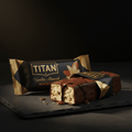 A chocolate-drizzled TITAN Vanilla Almond protein bar on a slate surface, next to its black and gold wrapper.