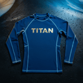 Blue long-sleeve TITAN rashguard with gold logo, crew neck, and contrast stitching on a wet, dark background.